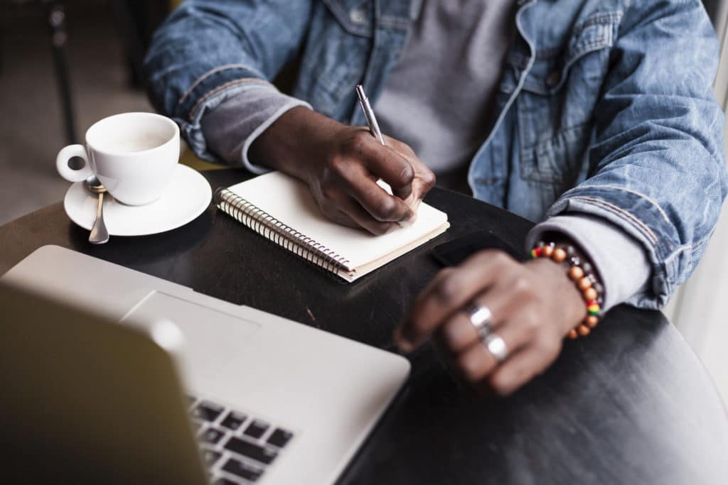 Un homme assis à son bureau, écrit dans un carnet à côté d'un ordinateur portable et d'une tasse de café, symbolisant le processus de rédaction professionnelle